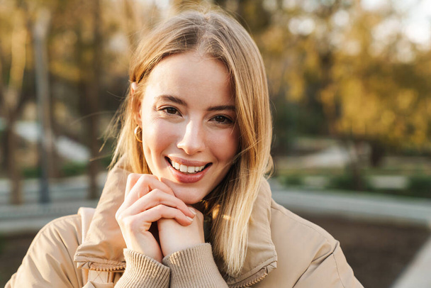 Portrait of young blonde woman wearing jacket smiling and looking at camera while walking in park - 写真・画像