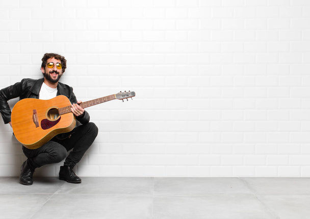 young musician man smiling happily with a hand on hip and confident, positive, proud and friendly attitude with a guitar, rock and roll concept - Фото, изображение