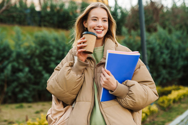 Portrait of smiling young woman holding exercise books and drinking coffee takeaway while walking in park - Zdjęcie, obraz