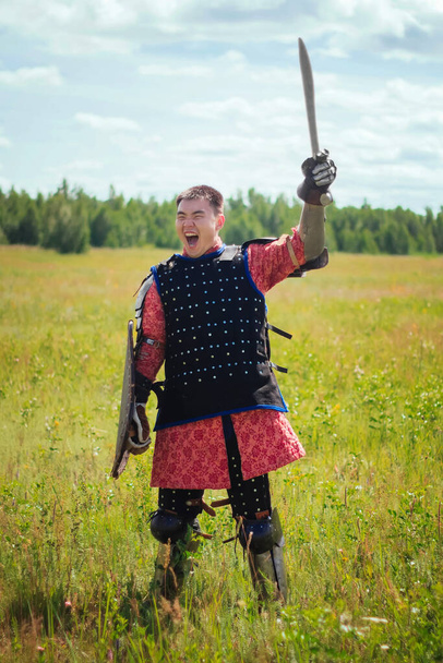 An Asian medieval warrior nomad in armor stands in a defensive position with a shield and a sword in his hands in the steppe against the background of the forest. Concept art battle in the field. - Fotó, kép