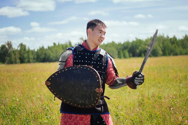 An Asian medieval warrior nomad in armor stands in a defensive position with a shield and a sword in his hands in the steppe against the background of the forest. Concept art battle in the field. - Fotoğraf, Görsel