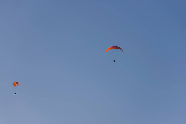 Paragliders with multi-colored parachutes fly high in the clear blue sky over steep cliffs. Parachuting, adrenalitis, love of height, risk, freedom of action. Desire to be able to fly - Photo, image