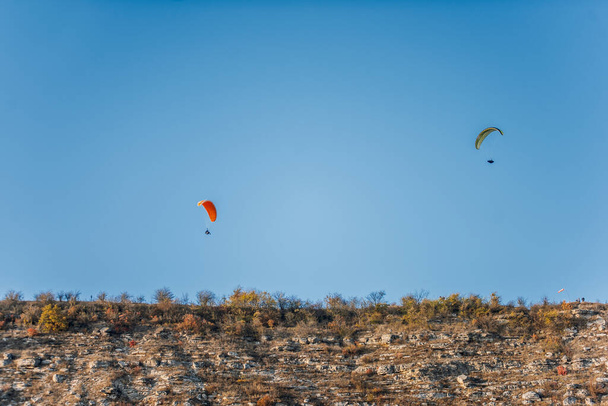 Paragliders with multi-colored parachutes fly high in the clear blue sky over steep cliffs. Parachuting, adrenalitis, love of height, risk, freedom of action. Desire to be able to fly - Foto, imagen