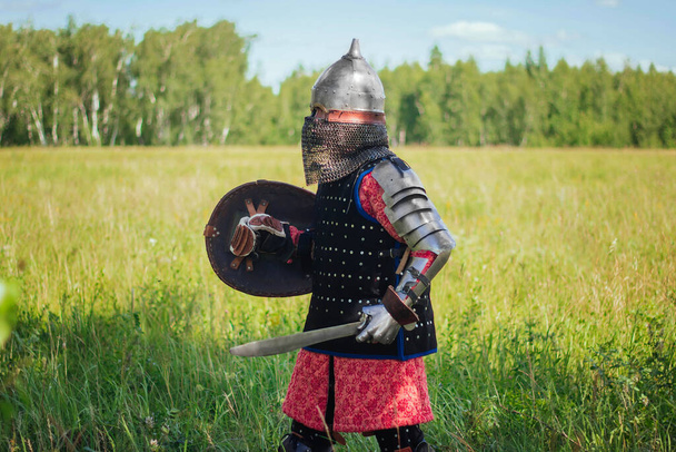 A medieval Central Asian warrior, a nomad in 14th-century armor, stands in a defensive or attacking position with a shield and a saber in his hands in a field against a forest. - 写真・画像
