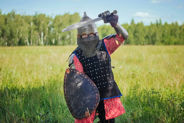 A medieval Central Asian warrior, a nomad in 14th-century armor, stands in a defensive or attacking position with a shield and a saber in his hands in a field against a forest. - Zdjęcie, obraz