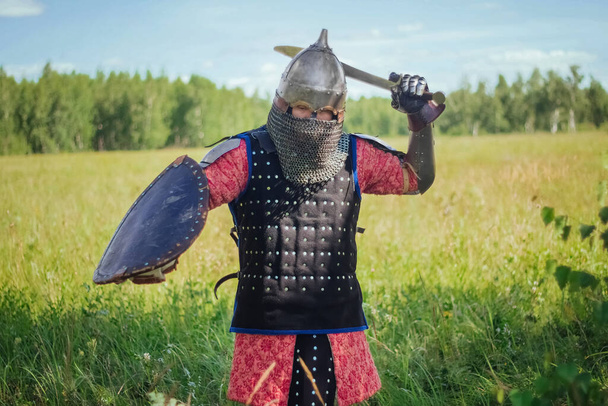 A medieval Central Asian warrior, a nomad in 14th-century armor, stands in a defensive or attacking position with a shield and a saber in his hands in a field against a forest. - Photo, image
