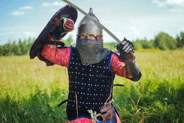 A medieval Central Asian warrior, a nomad in 14th-century armor, stands in a defensive or attacking position with a shield and a saber in his hands in a field against a forest. - Foto, immagini