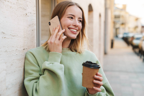 Portrait of smiling young woman drinking coffee takeaway and talking on mobile phone while walking at city street - Photo, image