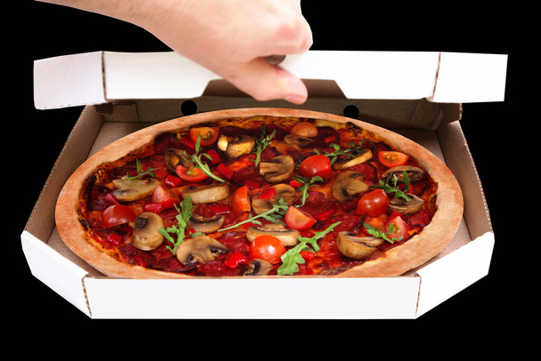 Um homem abre a caixa de papelão, serviço de entrega de pizza. Deliciosa pizza quente com salame picante, rúcula, tomate cereja, cogumelos e mistura de especiarias texas, sobre fundo preto
 - Foto, Imagem