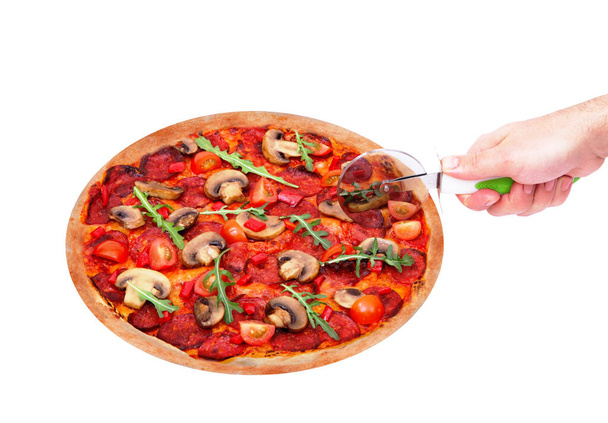 La main du chef coupe la délicieuse pizza chaude avec salami épicé, roquette, tomates cerises, champignons et texas mélange d'épices, avec un coupe-pizza, isolé sur fond blanc, vue d'angle
 - Photo, image