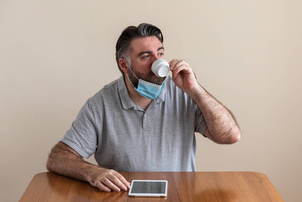 Un hombre lleva mascara para protegerse del covid19 coronavirus para no contagiarse. mientras trabaja a traves de ordenador y tableta. tiene una taza de café y esta en videoconferencia. con barba
. - Foto, Imagen