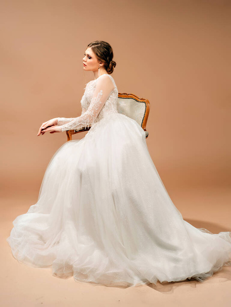 Красивая невеста в белом свадебном платье сидит в винтажном кресле. Студийный портрет на коричневом фоне. Модное свадебное платье
.  - Фото, изображение