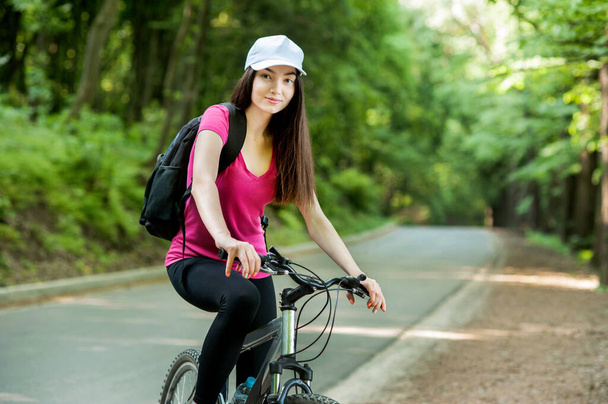 ragazza ciclista bruna con cappuccio bianco su una bicicletta su strada asfaltata nella foresta
 - Foto, immagini
