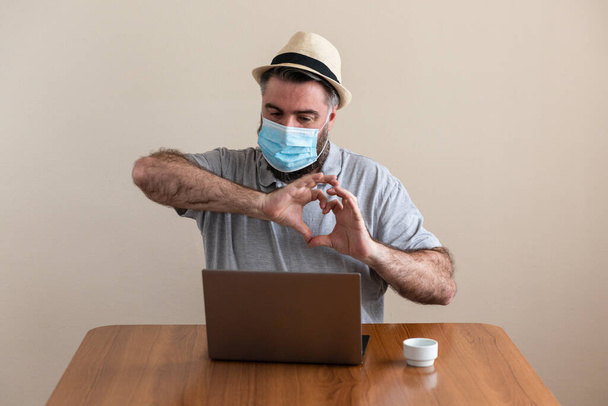 Un hombre lleva mascarilla para protegerse del covid19 coronavirus para no contagiarse. mientras trabaja a travs de ordenador y tableta. Videoconferencia. el hombre lleva barba, caucsico y sombrero. hace el smbolo del corazn con las manos
 - Foto, Imagen