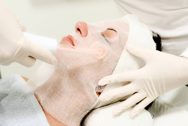 Kozmetikus fehér kesztyűben kezeli a női ügyfél elektromos darsonval arcmasszázs, eljárás szépségszalonban - Fotó, kép
