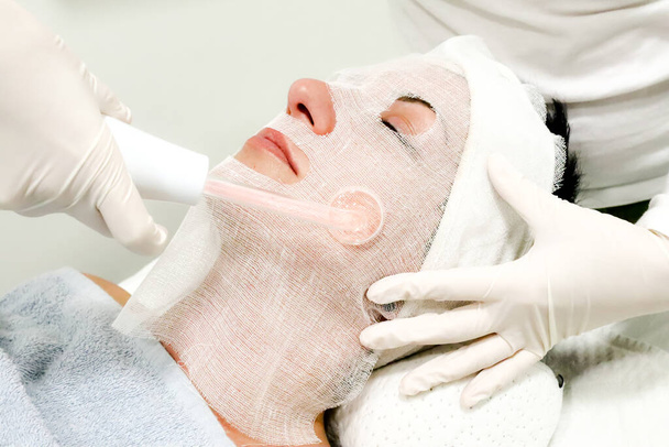 Beyaz eldivenli güzellik uzmanı bayan müşteriye elektrikli Darsonval yüz masajı ve güzellik salonunda işlem uyguluyor. - Fotoğraf, Görsel