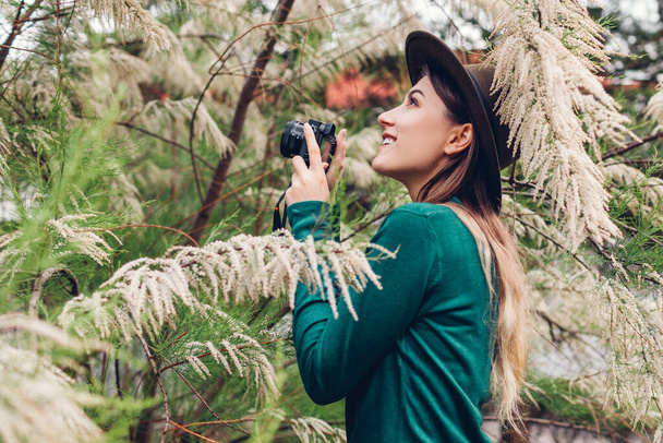 Νεαρή κομψή γυναίκα τραβά φωτογραφίες χρησιμοποιώντας compact φωτογραφική μηχανή στον καλοκαιρινό κήπο. Κορίτσι απολαμβάνοντας χόμπι στο πάρκο γυρίσματα ανθισμένα δέντρα - Φωτογραφία, εικόνα