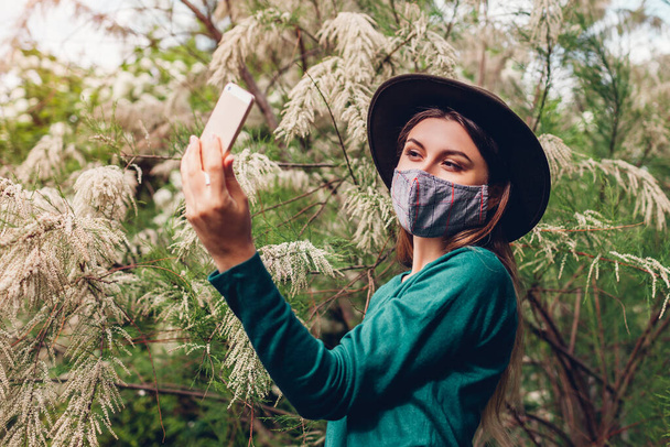 Chica tomando selfie en el teléfono inteligente en el jardín en flor. Mujer lleva máscara protectora al aire libre durante coronavirus covid-19 pandemia
. - Foto, Imagen