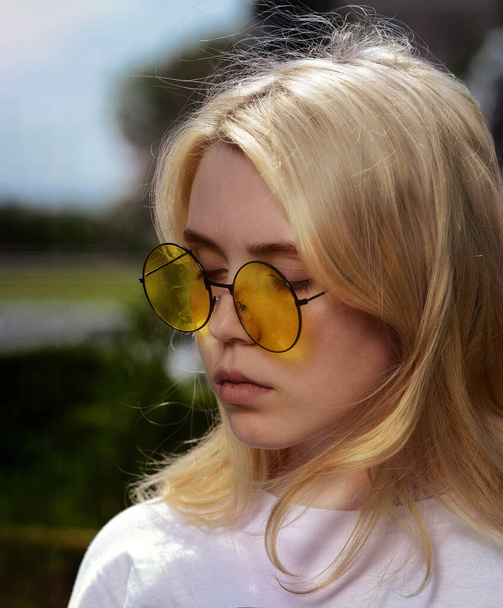 A young blonde girl outdoors in summer in yellow round glasses closed her eyes thoughtfully. Soft focus - Foto, afbeelding