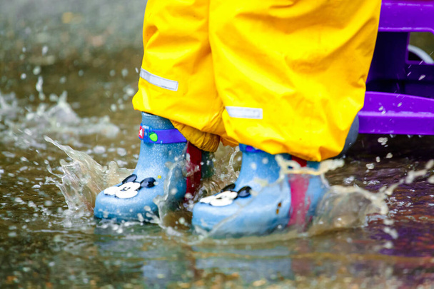 babyvoeten in blauwe rubberen laarzen en gele broek. spray vanaf de sprong. na de regen. - Foto, afbeelding
