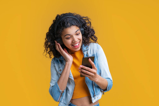 Linda chica de piel oscura sonriente en auriculares inalámbricos disfrutando escuchando música utilizando la aplicación en su teléfono móvil
. - Foto, imagen