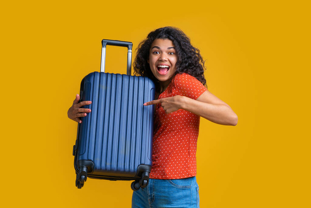 Волнующая темнокожая девушка с темно-синим ручным багажом и указывающая указательным пальцем на сумку. Студия снята на желтом фоне
. - Фото, изображение