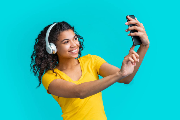 Okouzlující mladá žena v bezdrátových sluchátkách poslouchat hudbu a brát selfie na svém mobilním telefonu. Roztomilá dívka s dokonalým úsměvem s videokonferenci se svým přítelem. - Fotografie, Obrázek