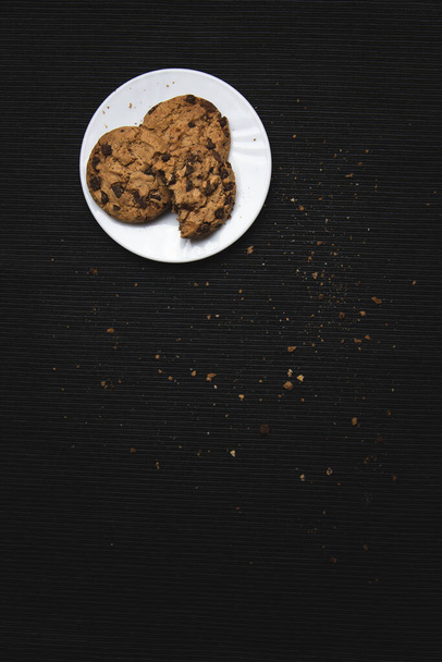 Chocolade chip koekjes in een witte plaat, op zwarte stof achtergrond vol met kruimels en chocolade chips. Ongezond eten, heerlijke traktatie, calorierijk voedsel - Foto, afbeelding