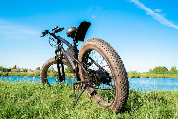 Чорний електричний велосипед з товстими колесами на траві біля озера. Фатбайк
. - Фото, зображення