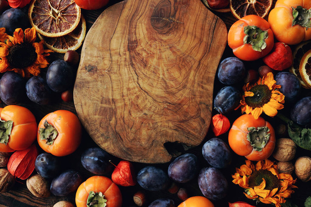 Autumn still-life composition with fruits and vegetables. Assortment of plums, persimmon, walnut, sunflowers and dry oranges and rustic wooden board, top view, space for text - Foto, afbeelding