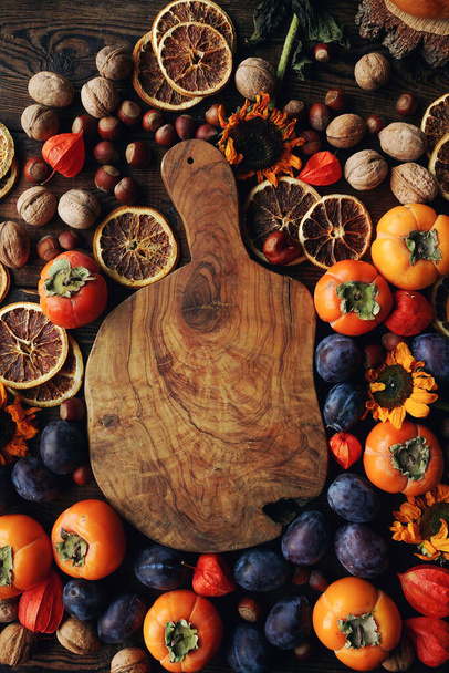 Autumn still-life composition with fruits and vegetables. Assortment of plums, persimmon, walnut, sunflowers and dry oranges and rustic wooden board, top view, space for text - Fotoğraf, Görsel