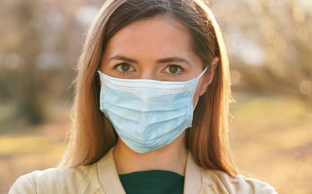 Young woman wearing blue disposable virus mouth nose mask, nice sunny bokeh in background, closeup face portrait - Foto, Imagem