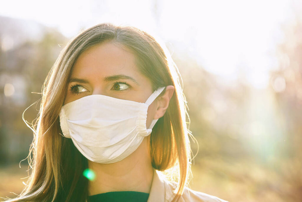 Young woman wearing white cotton virus mouth nose mask, nice backlight sun bokeh in background, closeup face portrait - Foto, immagini