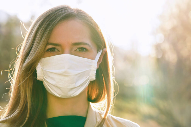 Young woman wearing white cotton virus mouth nose mask, nice backlight sun bokeh in background, closeup face portrait - Foto, Imagem
