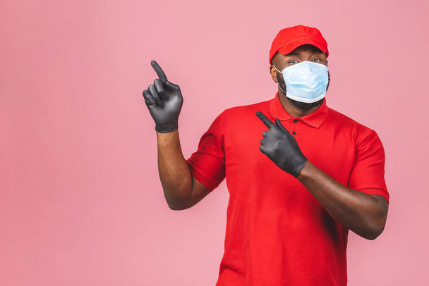 Liefermann in roter Mütze leeres T-Shirt einheitliche sterile Gesichtsmaske Handschuhe isoliert auf rosa Hintergrund. Guy Mitarbeiter arbeitet Kurierdienst Quarantäne-Pandemie Coronavirus. Zeigefinger.  - Foto, Bild