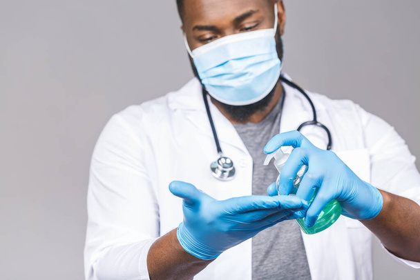 灰色の背景に隔離された顔マスク手袋のアフリカ系アメリカ人医師の男性。流行性大流行性コロナウイルス2019-nconvは、 covid-19インフルエンザウイルスを発見しました。瓶液抗菌消毒剤の保持. - 写真・画像