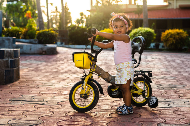 Παιχνιδιάρικο Όμορφο Ινδικό κορίτσι παιδί / νήπιο / νήπιο που φοράει μια κορδέλα μαλλιών, παίζει με ένα ποδήλατο / τρίκυκλο. Το παιδί δίνει χαρούμενες εκφράσεις - Φωτογραφία, εικόνα