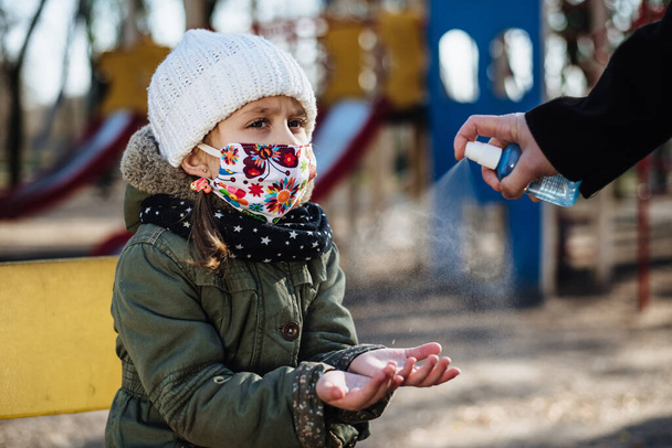 公園の遊び場を背景に、母親は少女の手に抗菌スプレーを消毒します。Covid-19感染に対する予防措置。保護マスクの子供は屋外を歩く。フィルムノイズ - 写真・画像
