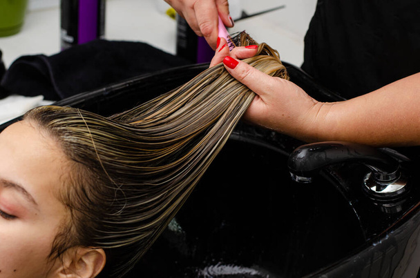 Peluquero aplicando mascarilla para el cabello y peinando el cabello húmedo con cepillo en un fregadero negro
. - Foto, imagen