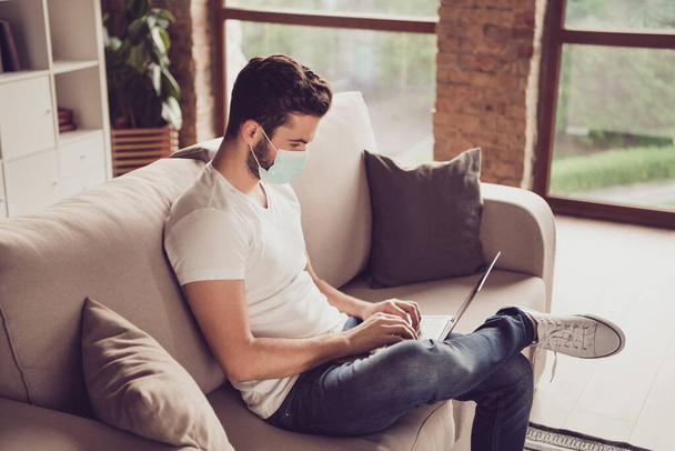 Profile photo of student young guy chatting notebook freelance job internet coach typing keyboard sms message quarantine stay home wear facial medical mask sit couch living room indoors - Foto, imagen