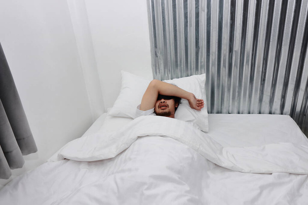 Νεαρός Ασιάτης με μάσκα ματιών που κοιμάται στο λευκό κρεβάτι στην κρεβατοκάμαρα. - Φωτογραφία, εικόνα
