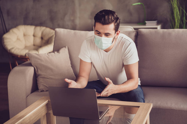 Photo of student young guy chatting notebook freelance job internet coach communicating employer want get job quarantine stay home wear facial medical mask sit couch living room indoors - 写真・画像