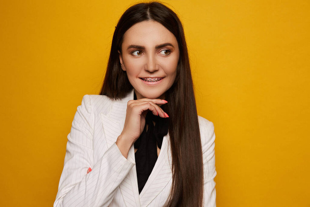 Portrait of a smiling young brunette woman with dental braces isolated at the yellow background. Model girl with gentle makeup and braces in a white jacket over yellow background. Dental business. - Foto, Bild
