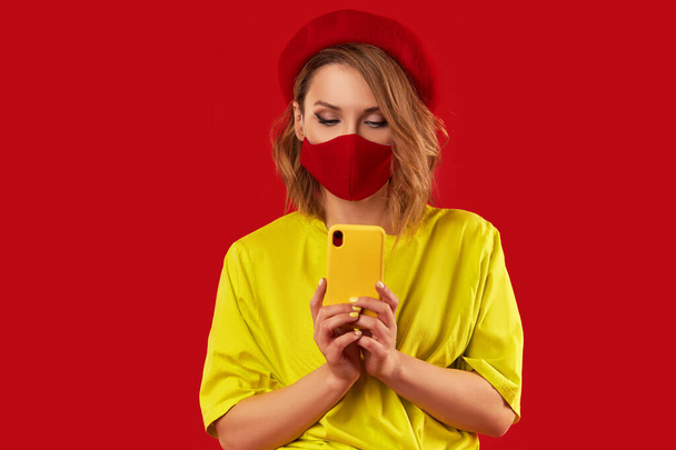 Woman in trendy fashion outfit during quarantine of coronavirus outbreak. Model in protective stylish face mask uses a smartphone on a red background - Фото, изображение
