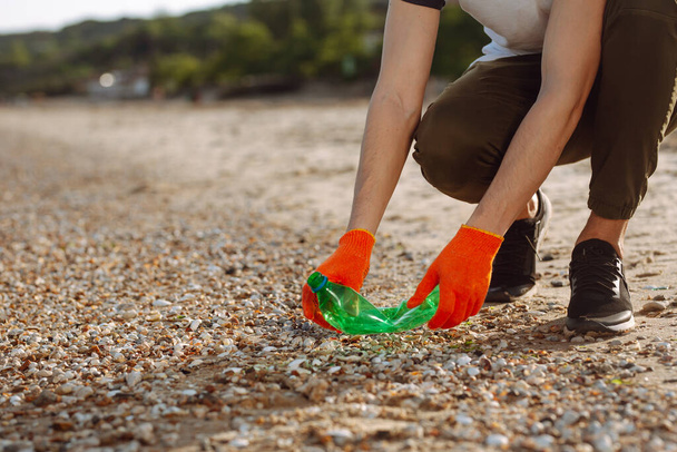 Young male volunteer picks up plastic garbage on the ocean coast. A man collects waste on the seaside to save ecology and protect environment. Recycle litter and pollution concept - Foto, Imagen