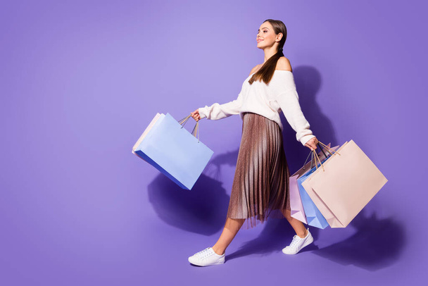 Full length profile side photo of positive satisfied girl enjoy shopping go walk copyspace store mall hold many bags wear brown sweater footwear isolated purple color background - Zdjęcie, obraz