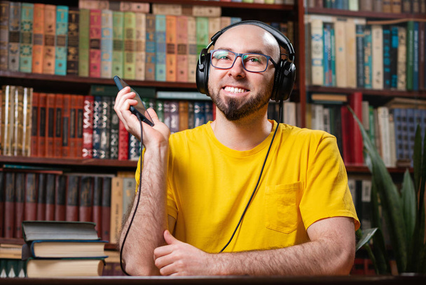 Egy kopasz férfi szemüveggel és szakállal, aki audiobookot vagy zenét hallgat fejhallgatóval. Könyvespolcok a háttérben. Az online szórakoztatás és a hangoskönyvek fogalma. - Fotó, kép
