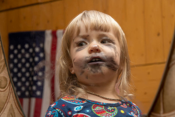Reflexe v zrcadle malé holčičky, která si namalovala obličej s dětskými značkami v očekávání prázdnin, můžete vidět americkou vlajku. Koncept: Den nezávislosti, první make-up, humor. - Fotografie, Obrázek