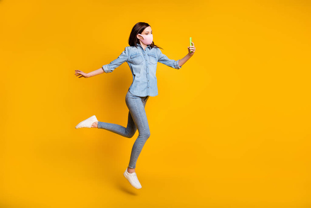 Full length photo of beautiful girl jump use smartphone make selfie video call connection technology wear medical mask denim jeans shirt isolated over yellow color background - Foto, Imagen