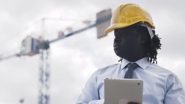 Portret Afrykanina w twardej czapce z tabletem na placu budowy i obserwacja prac - Materiał filmowy, wideo